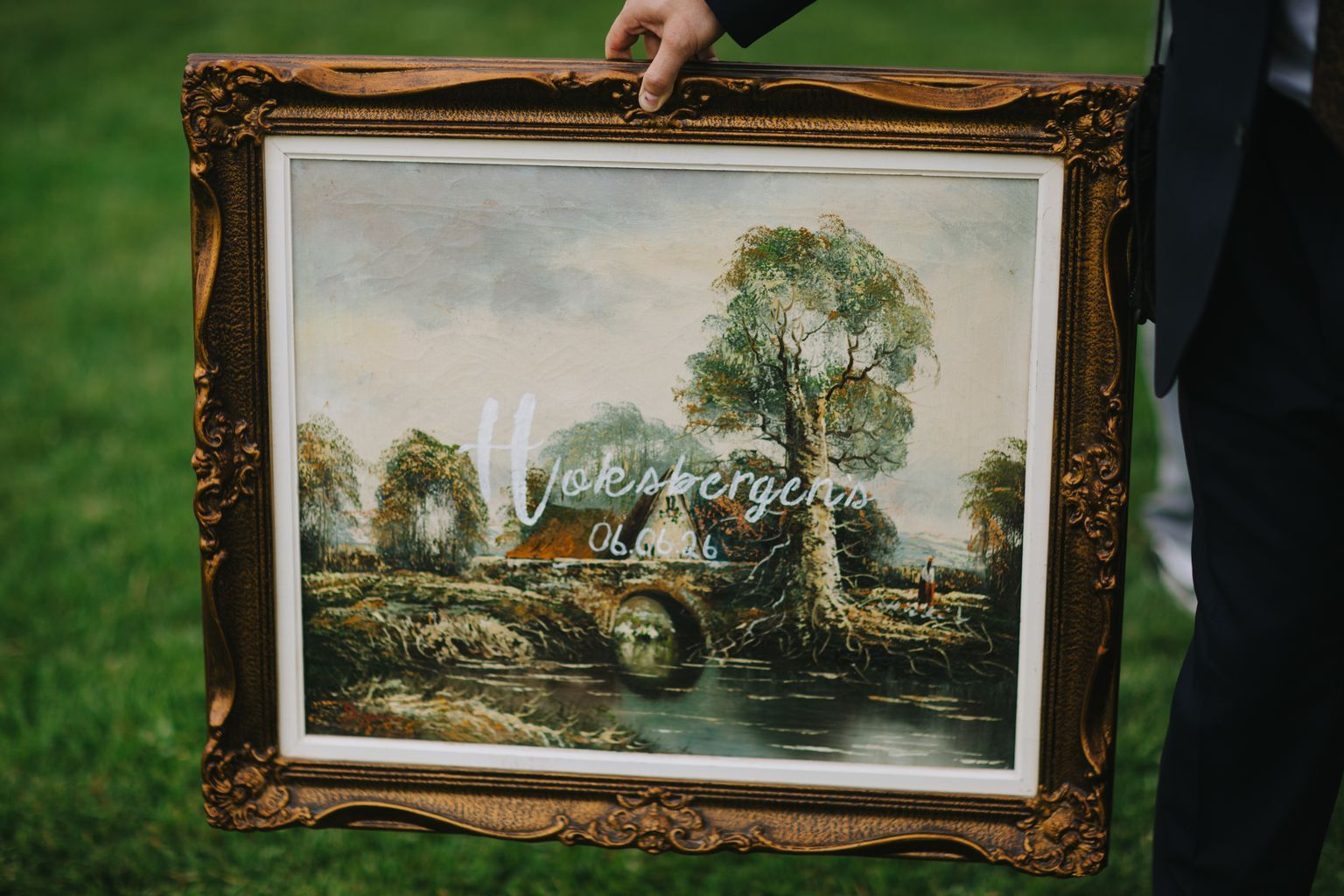 Hand holding ornate gold-framed landscape painting with "love and laughter" calligraphy overlay and wedding date