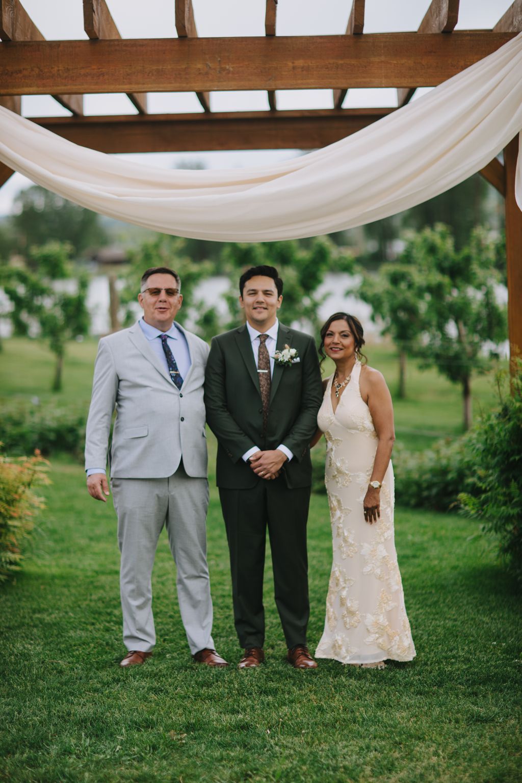 Groom in dark suit with boutonniere stands between two guests under wooden pergola with sheer draping on manicured lawn