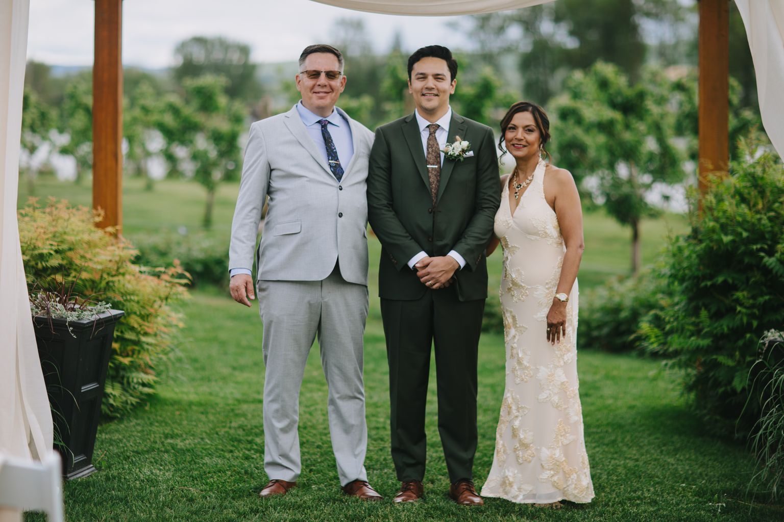 Groom in dark suit with boutonniere stands between two guests on grassy lawn during outdoor wedding ceremony