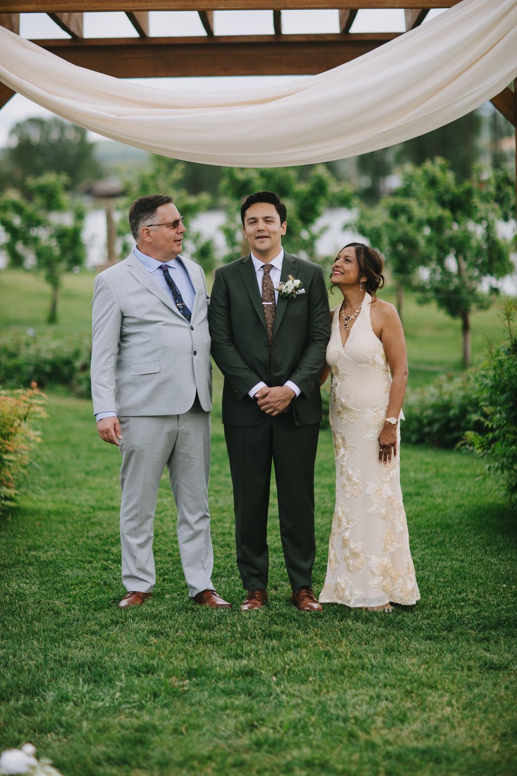 Bride in beaded white gown and groom in dark suit stand with officiant under wooden pergola with draped fabric on manicured lawn