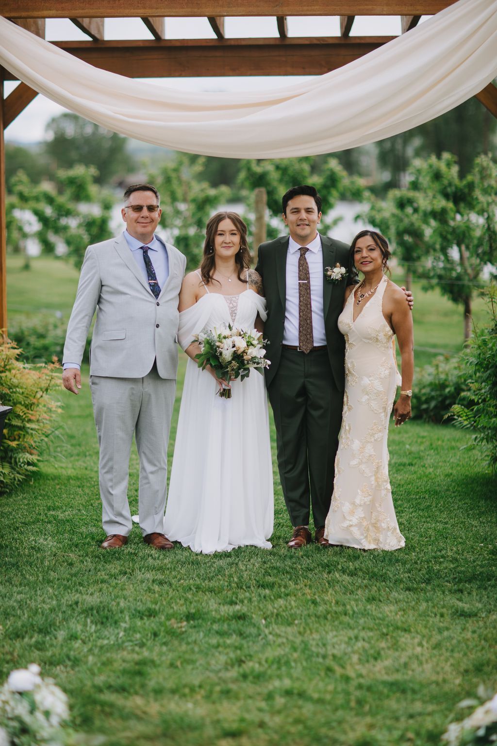 Bride in white gown and groom with family under wooden pergola with sheer draping on manicured lawn