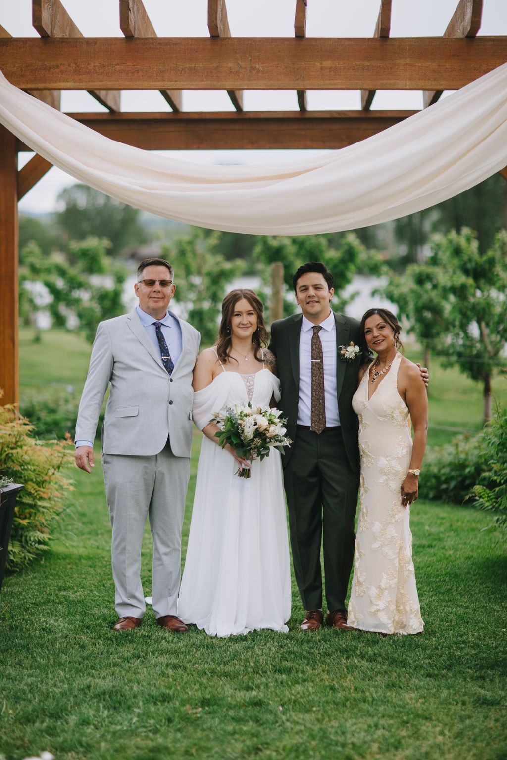 Bride in white dress holding white bouquet stands with groom and family under wooden pergola with sheer fabric draping