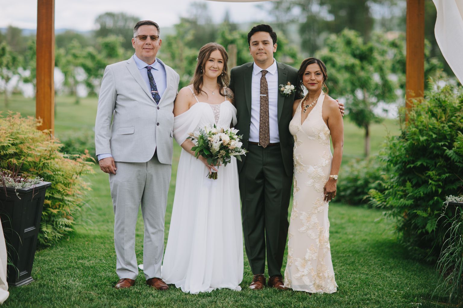 Bride in white dress holding white bouquet stands with groom in dark suit and family members on grass, mountain backdrop
