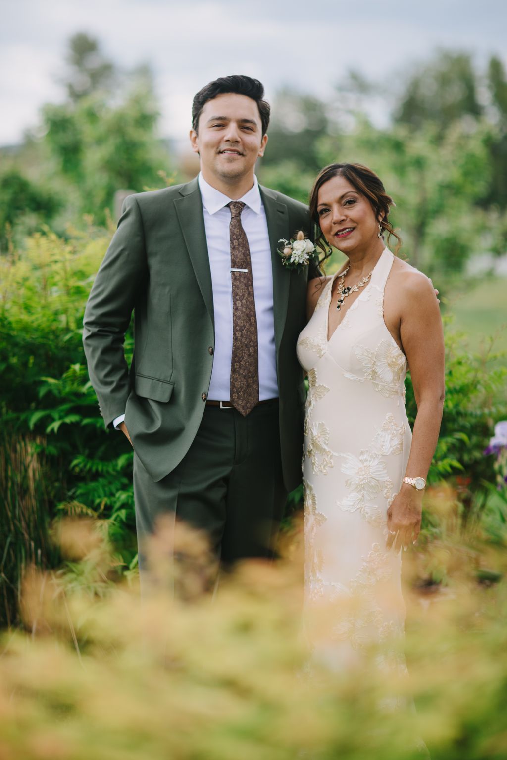 Groom in sage green suit and bride in cream embellished gown smile together in a sunlit garden setting