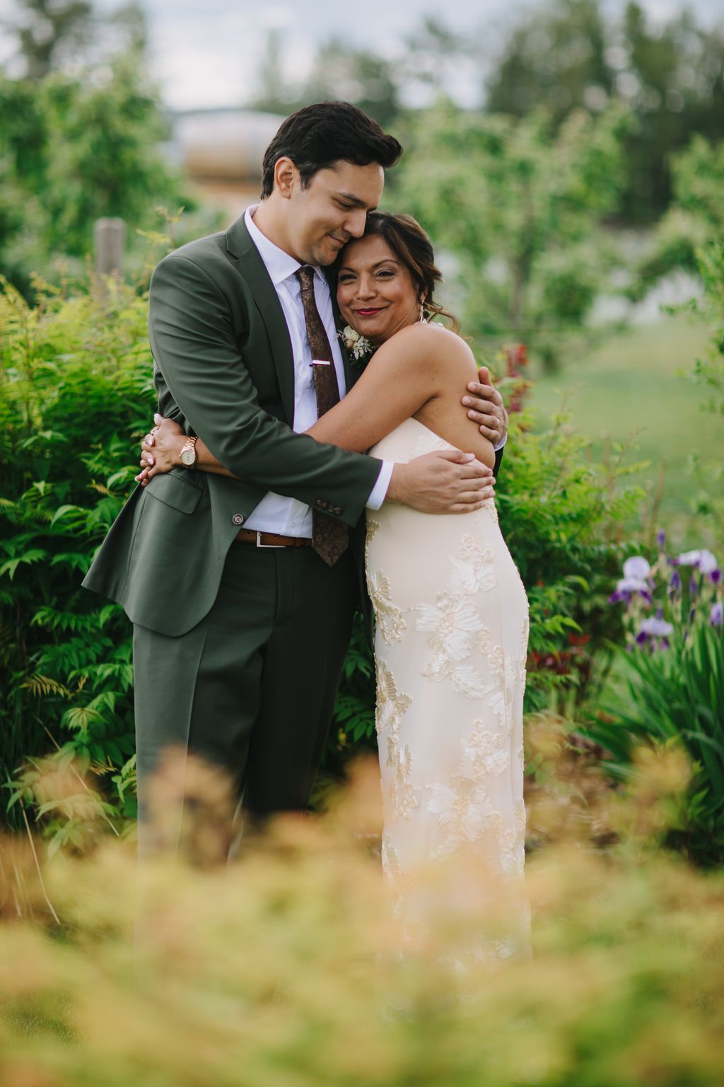 Groom in sage green suit embraces bride in ivory lace gown in a lush garden setting during golden hour