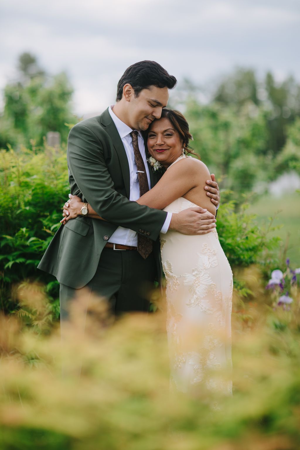 Groom in olive suit embraces bride in cream beaded gown outdoors with soft-focus green fields behind them