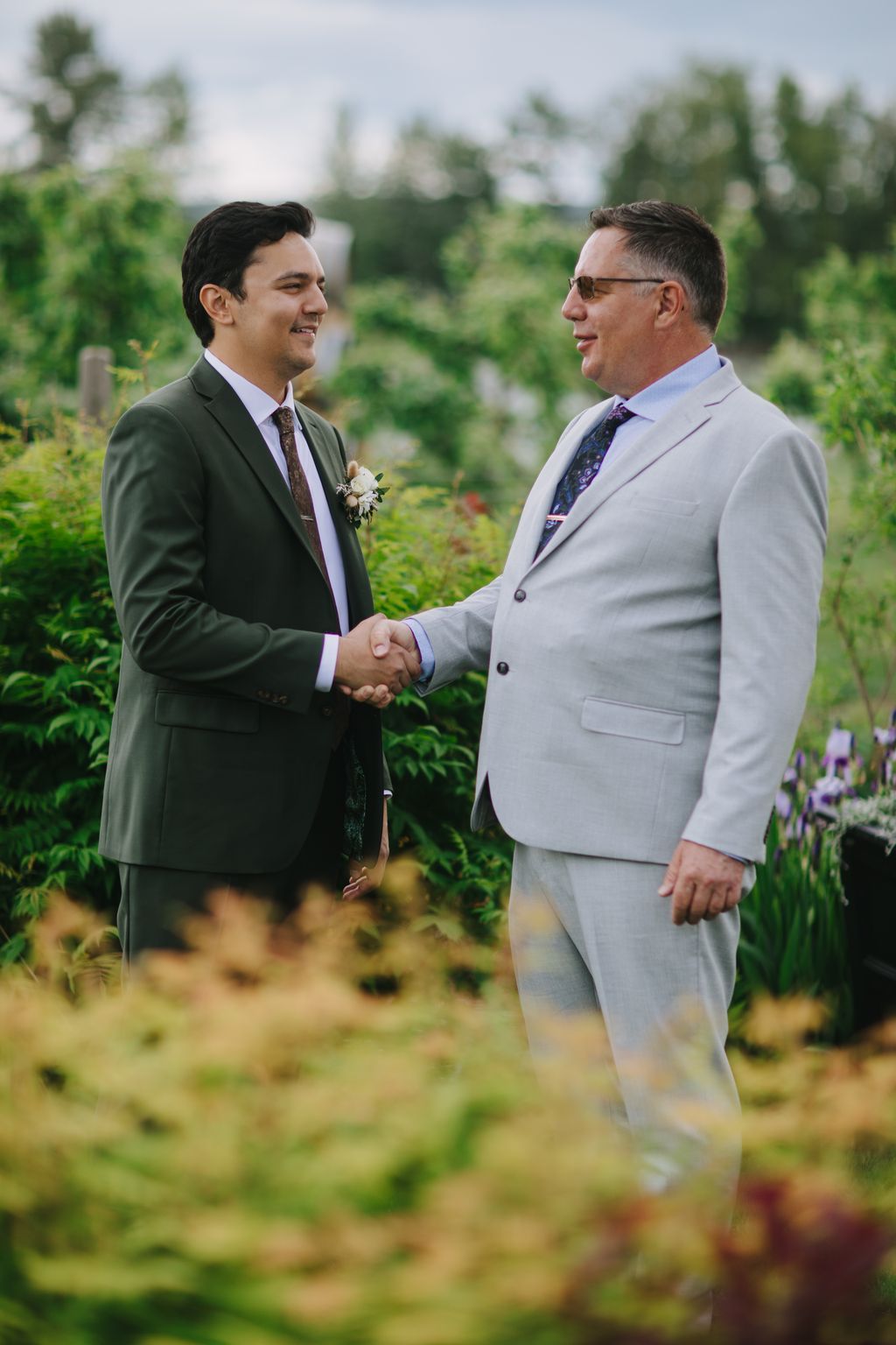 Two grooms in light suits shake hands and smile during their outdoor garden ceremony on an overcast day