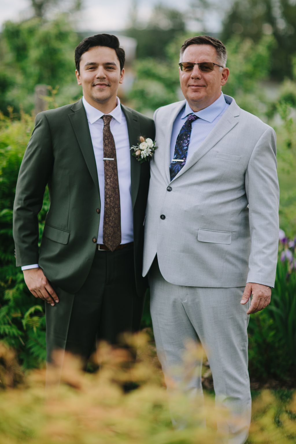 Two men in formal suits with boutonnières smile at the camera outdoors with soft, blurred green foliage behind them