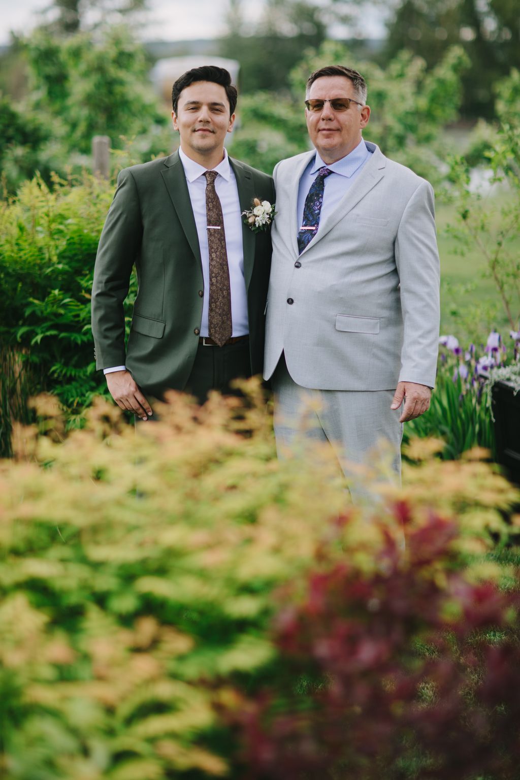 Two men in formal suits stand together outdoors with lush green foliage and water visible in the soft-focused background