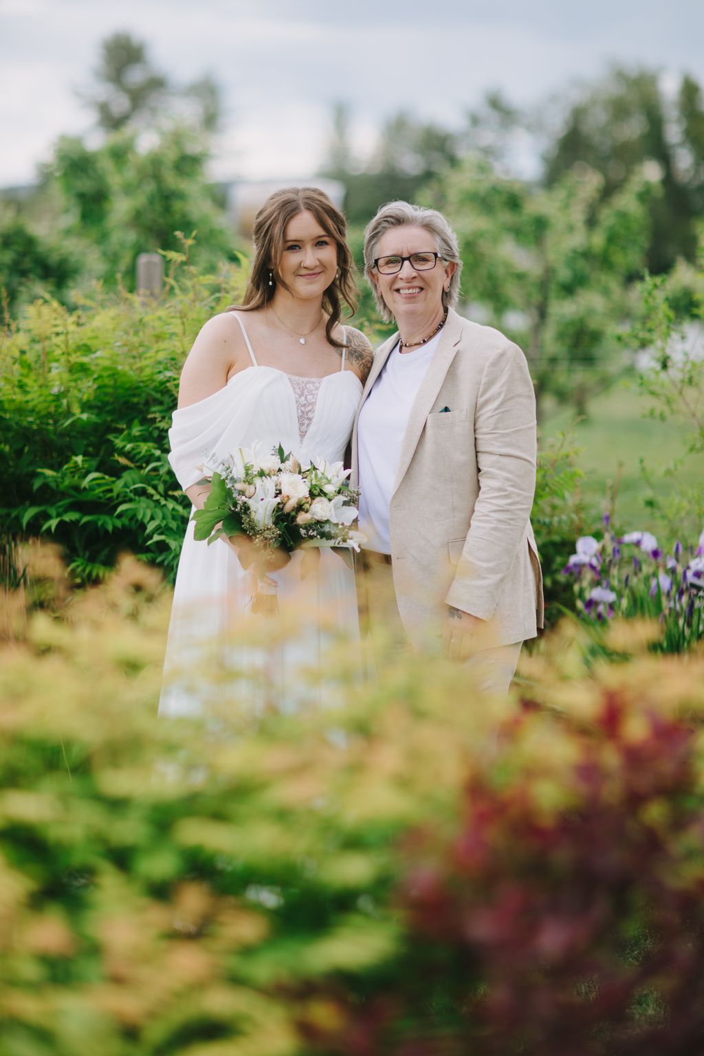 Bride in white gown and groom in beige suit smile together in a garden setting, holding a white flower bouquet