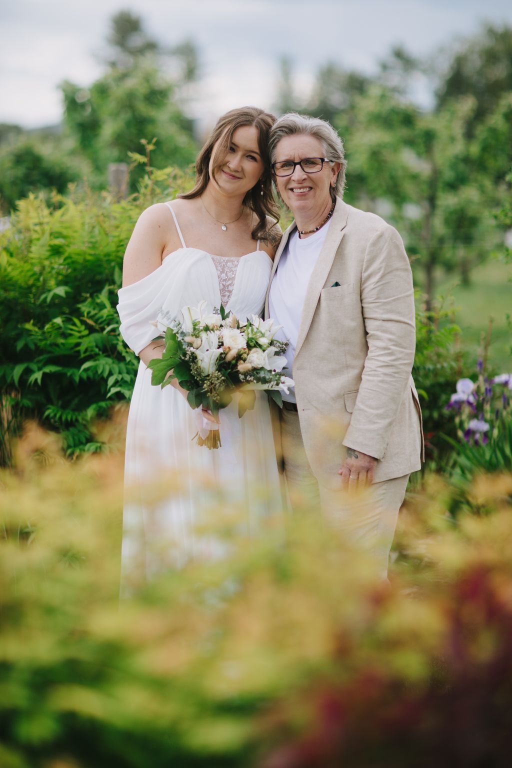 Bride in flowing white dress holding white bouquet smiles with older woman in beige blazer in sunlit garden setting