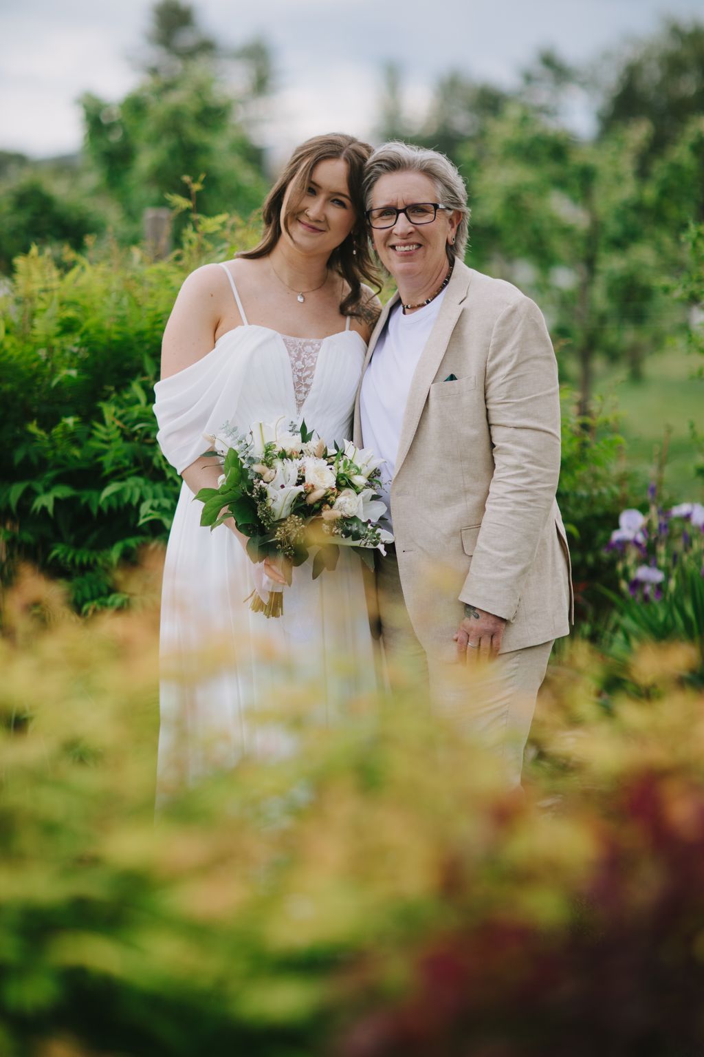 Two women smile together outdoors, one in a white wedding dress holding a white bouquet, the other in a beige blazer, with green garden foliage softly blurre…