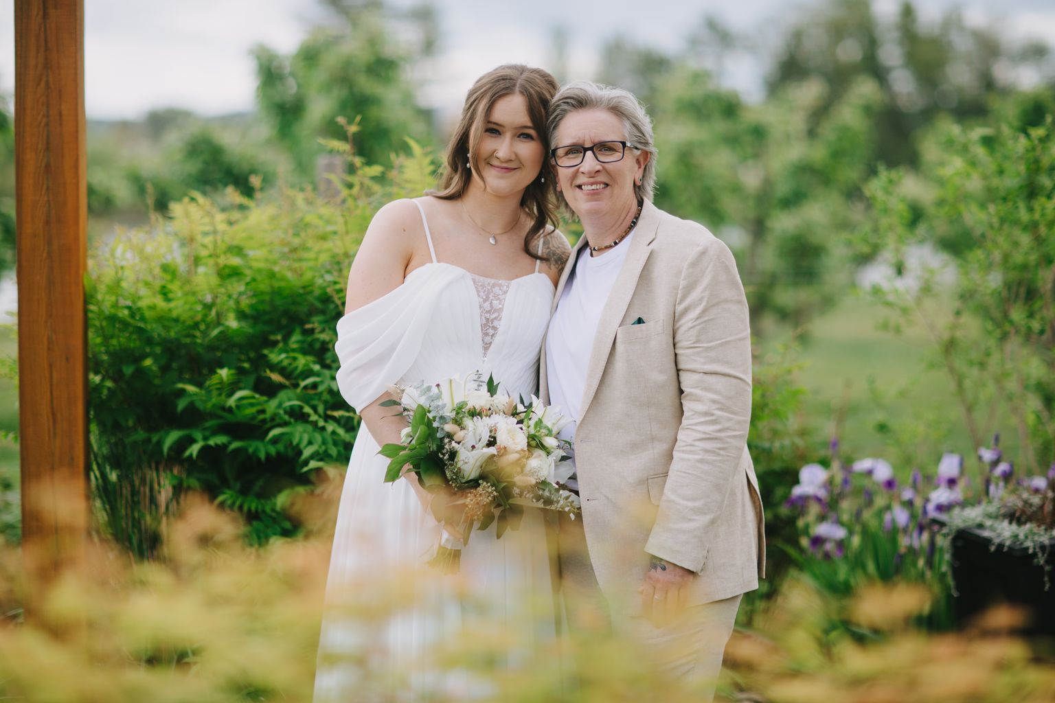 Bride in flowing white gown holding white flower bouquet stands smiling with partner in beige suit outdoors