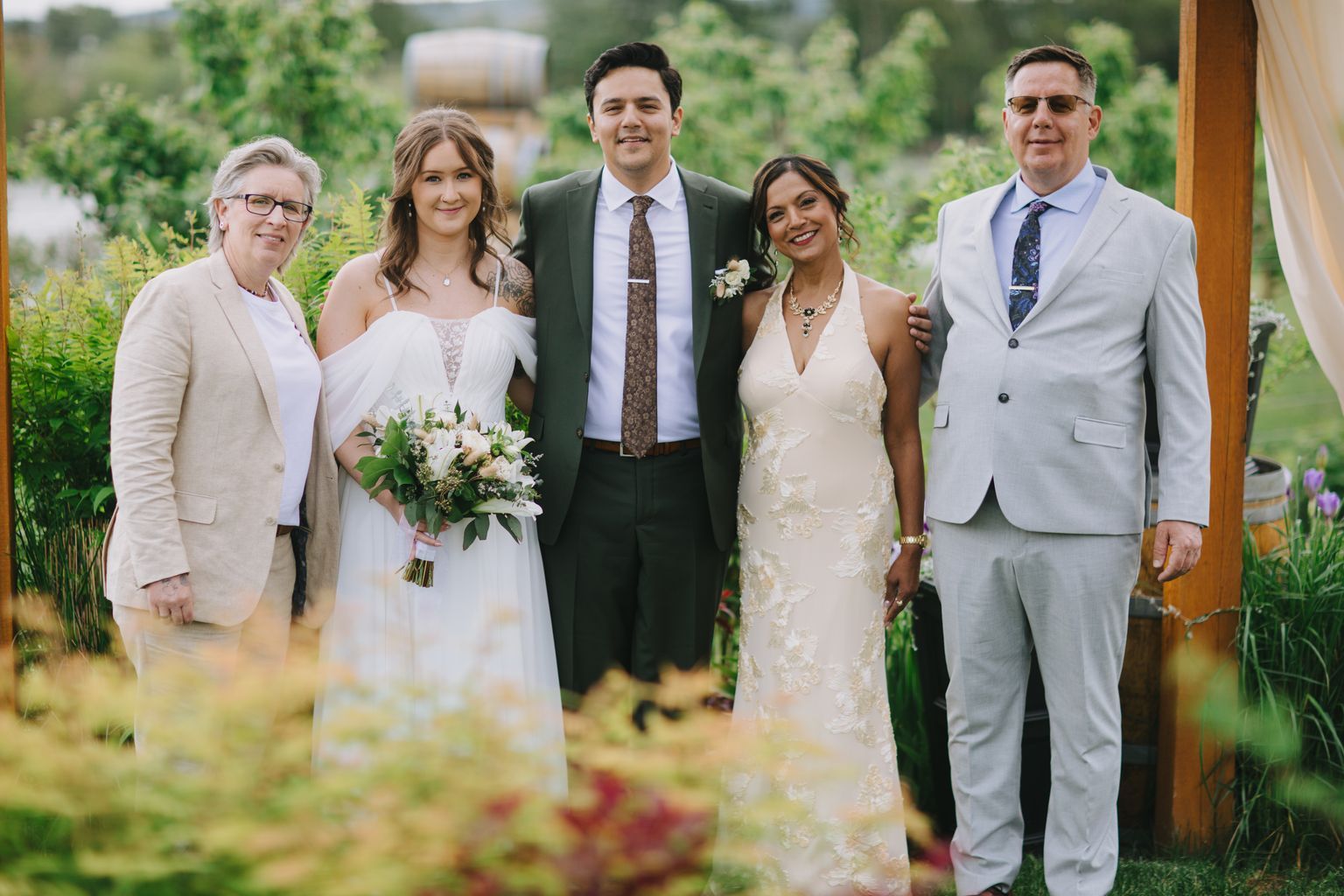 Groom in olive suit stands between two women in white dresses at outdoor garden wedding ceremony with green foliage background