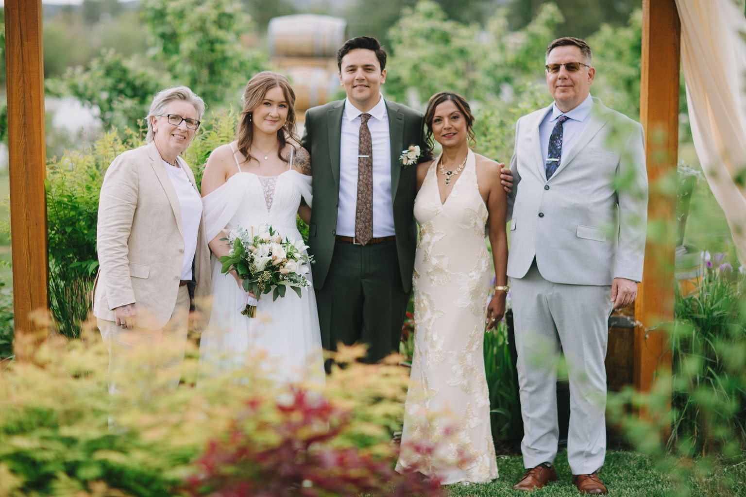 Bride in white lace dress and groom in olive suit with best man pose together in lush garden setting during daytime