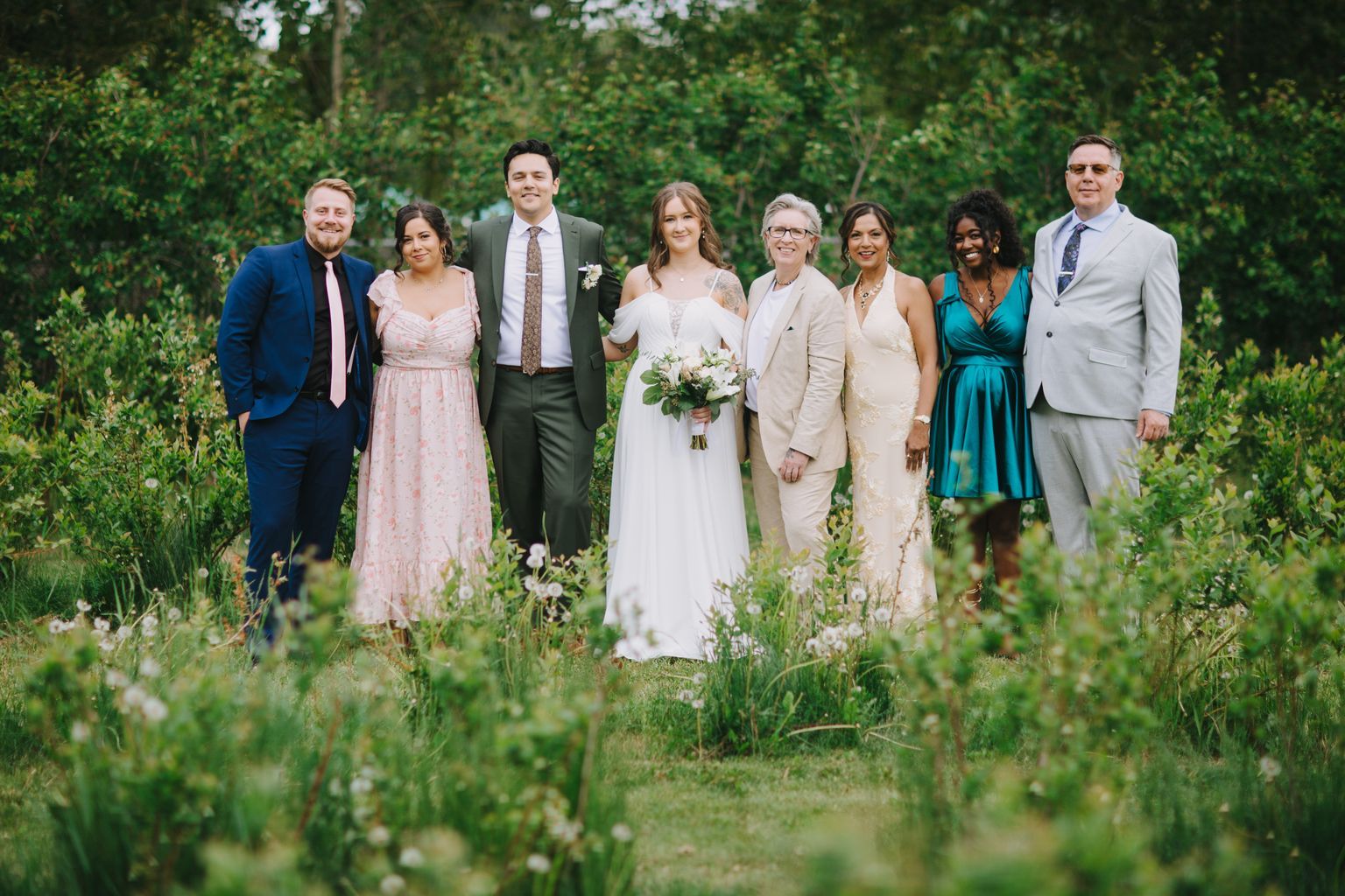 Bride in white dress holding bouquet stands with groom and family members in lush garden setting with green foliage background