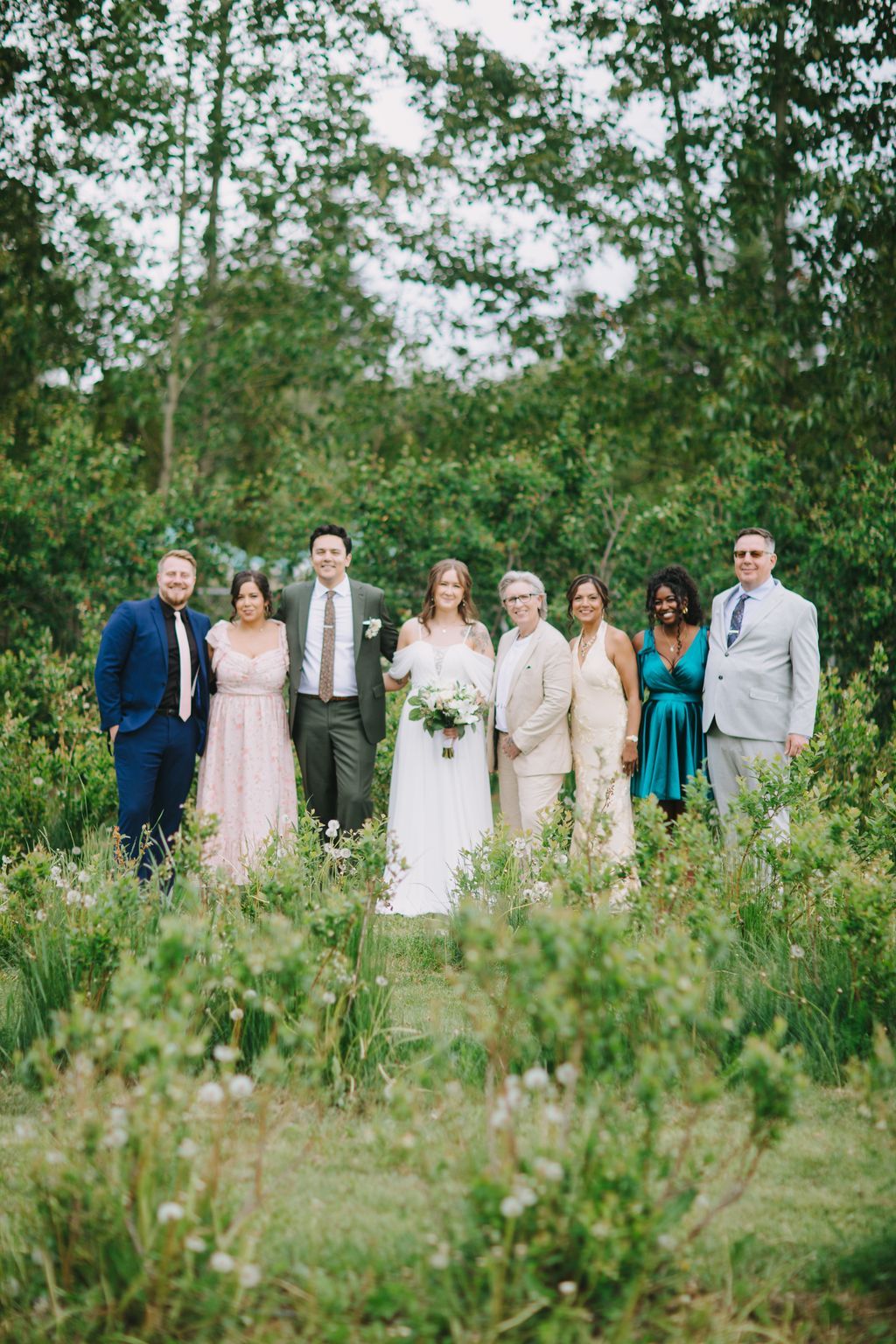 Bride in white dress with bouquet stands with groom and five family members in a wildflower field under leafy trees
