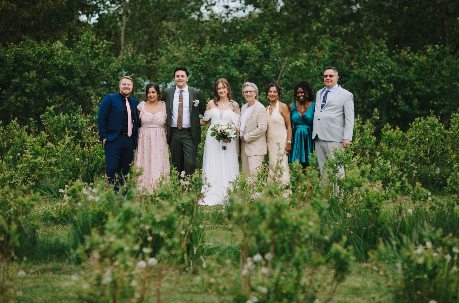 Bride in white dress and groom in green suit stand with wedding party among wildflowers and green foliage in natural outdoor garden setting
