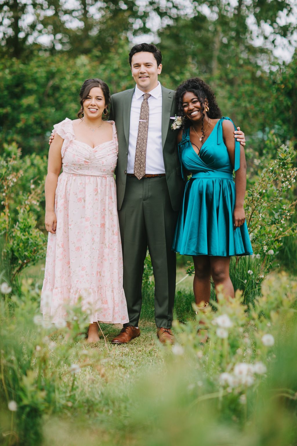 Groom in olive suit with two women in pink floral and teal dresses smiling outdoors with soft green foliage backdrop