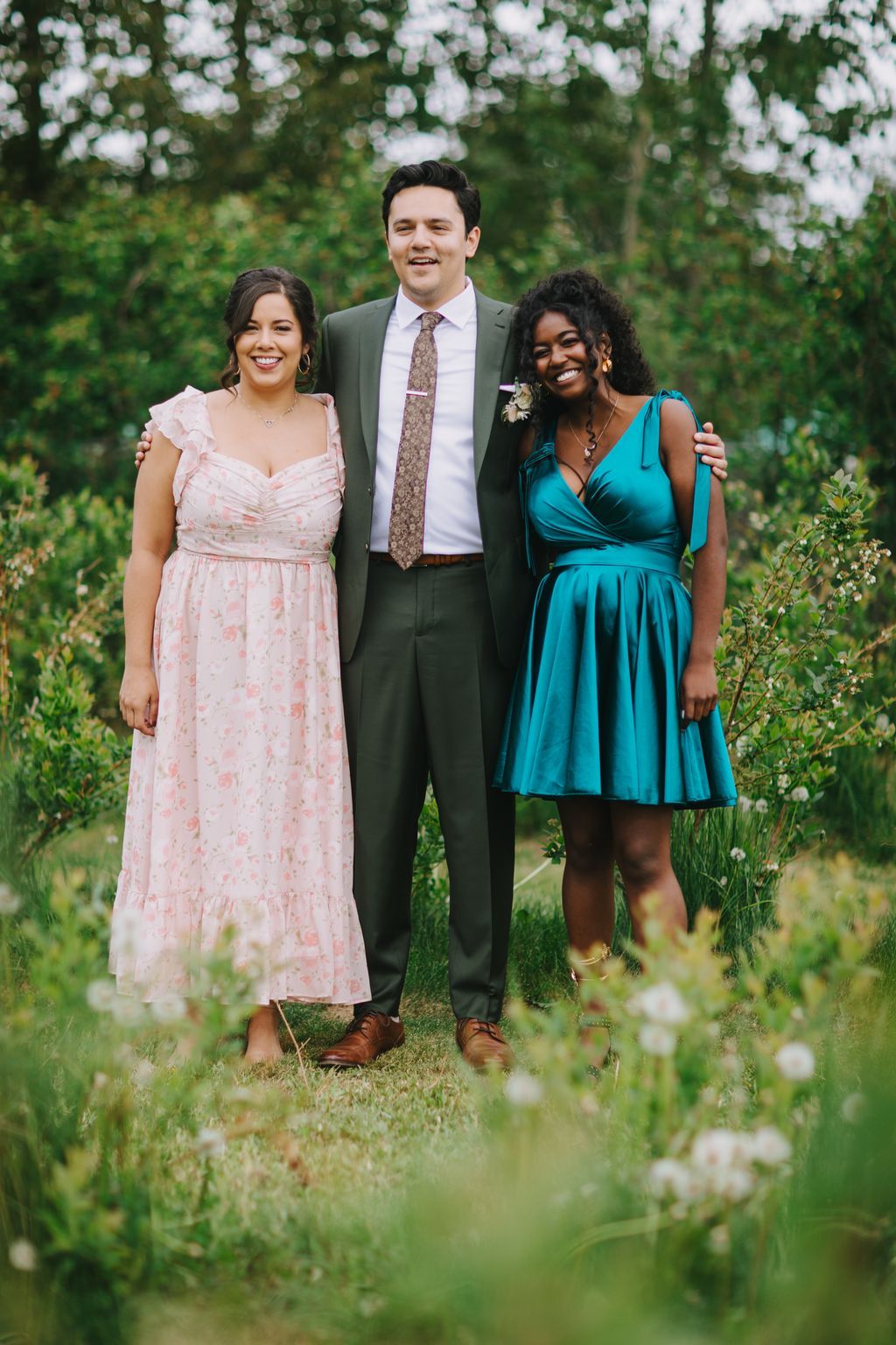 Three smiling guests stand together on grass with blooming trees and soft green foliage in the background