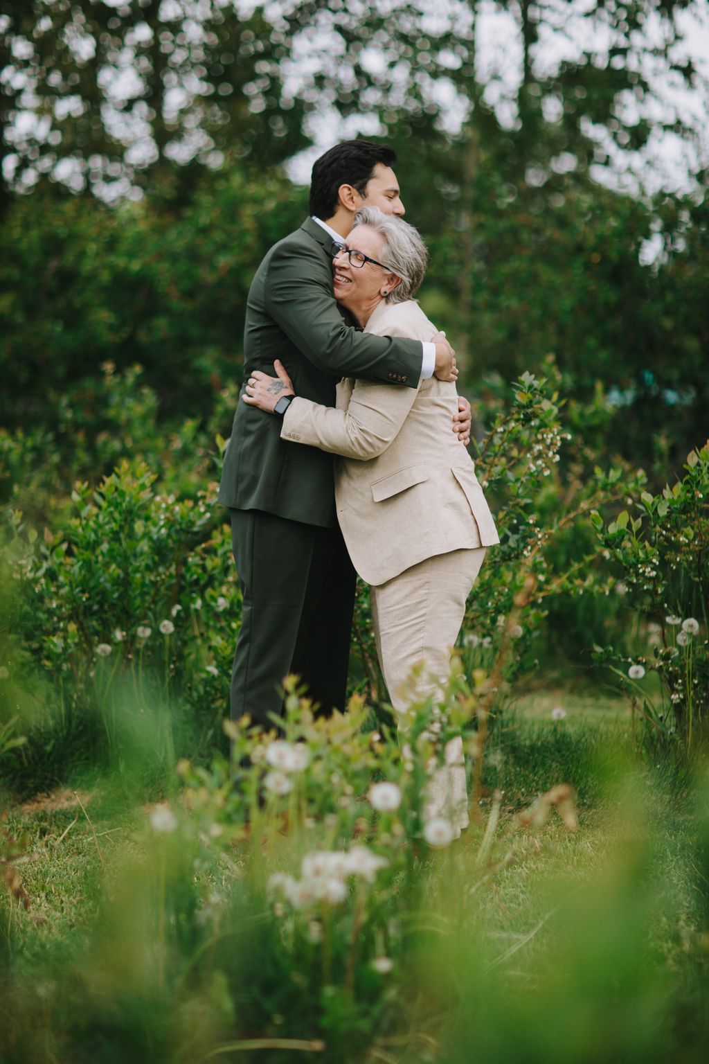 Two grooms embrace in a blooming garden, one in a sage green suit and the other in a cream suit, surrounded by white flowers and lush greenery
