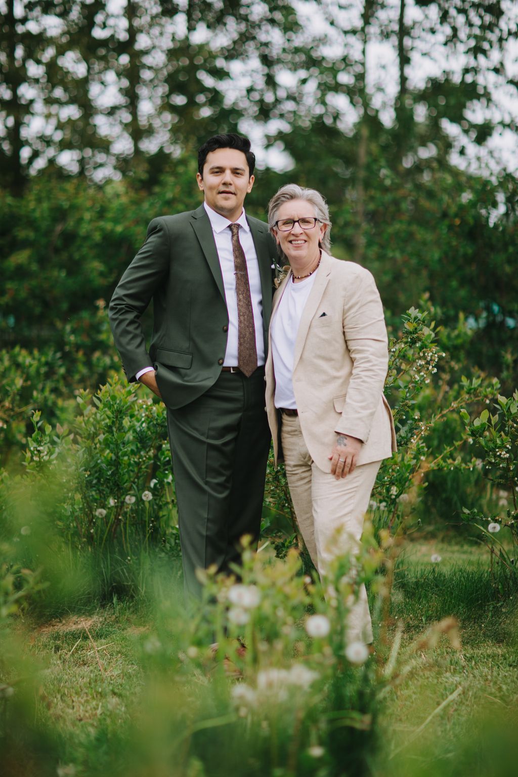 Two men in tailored suits stand smiling in a lush garden surrounded by white flowering trees and green foliage