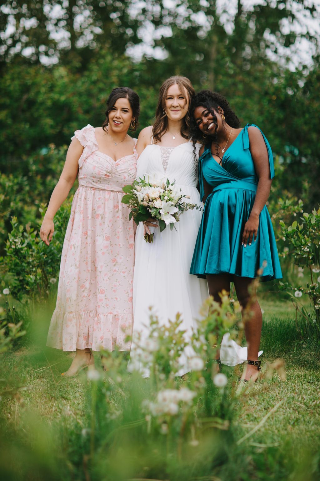 Bride in white dress with bouquet stands between two bridesmaids in pink and teal dresses in a lush garden setting