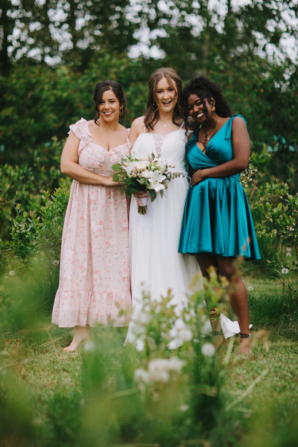 Bride in white dress with white bouquet smiling between two bridesmaids in pink and teal dresses in a lush garden setting