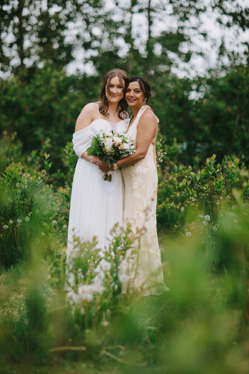 Two brides in white wedding dresses hold a bouquet surrounded by blooming white flowers in a lush garden