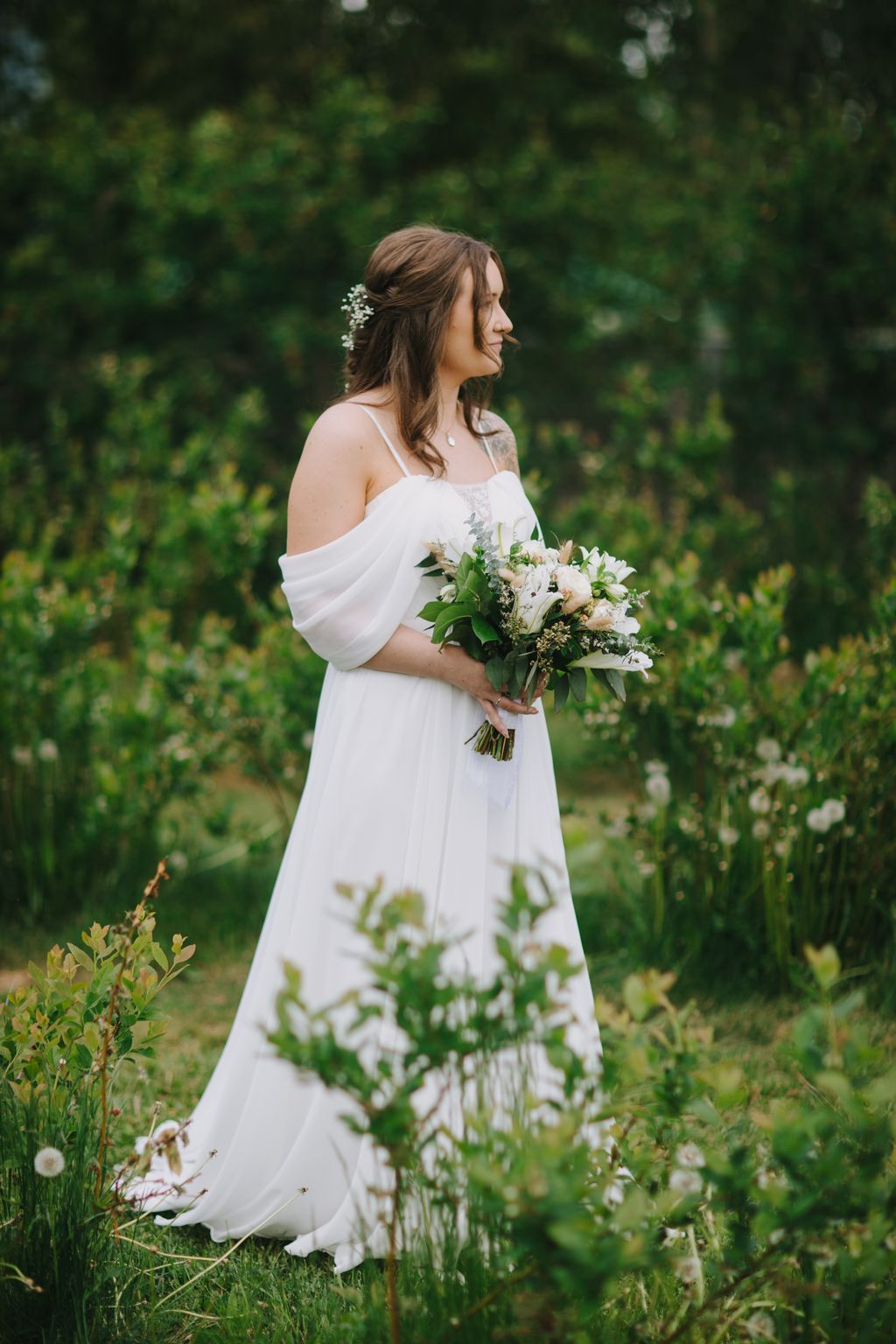 Bride in flowing white off-shoulder gown holds white and green bouquet, standing in lush garden with hair flowers and gentle smile