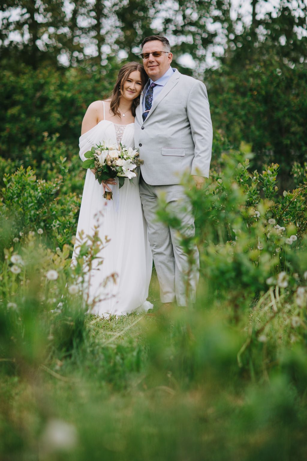 Bride in white off-shoulder gown and groom in light gray suit embrace outdoors surrounded by lush green foliage and flowering trees
