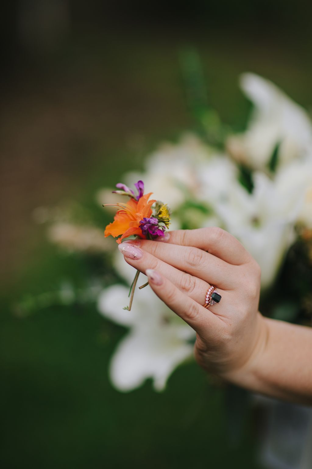 Bride''s hand with dark gemstone ring holding vibrant orange and purple flower against soft green garden backdrop