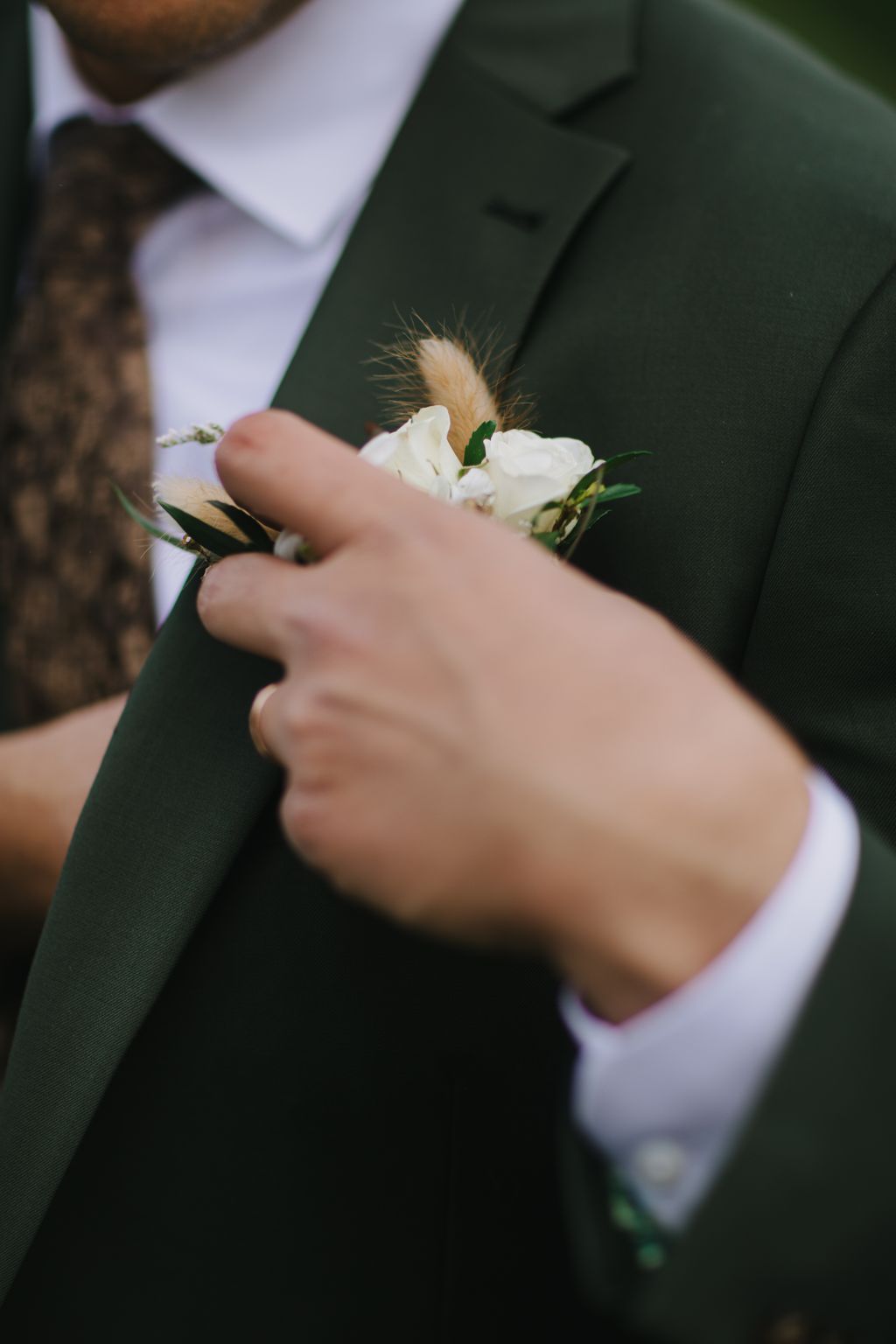 Groom in dark suit holding white rose boutonniere with feathered accent during wedding ceremony