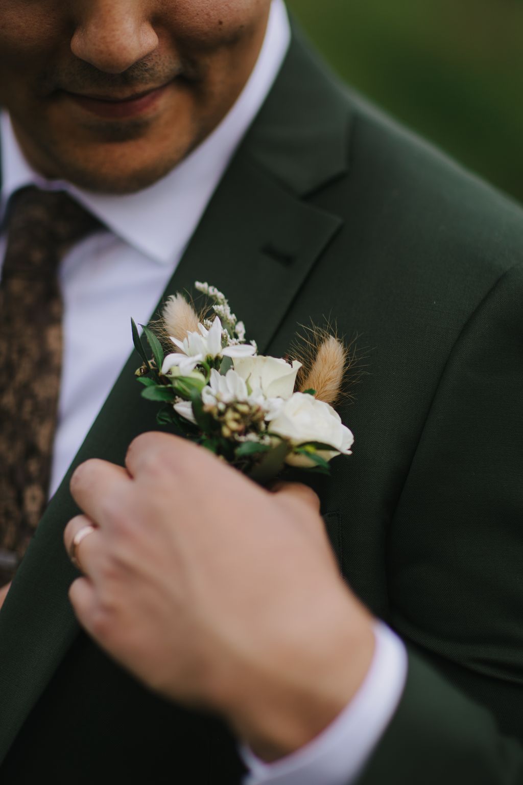 Groom in dark suit holding white and cream boutonniere with dried pampas grass detail against blurred green background