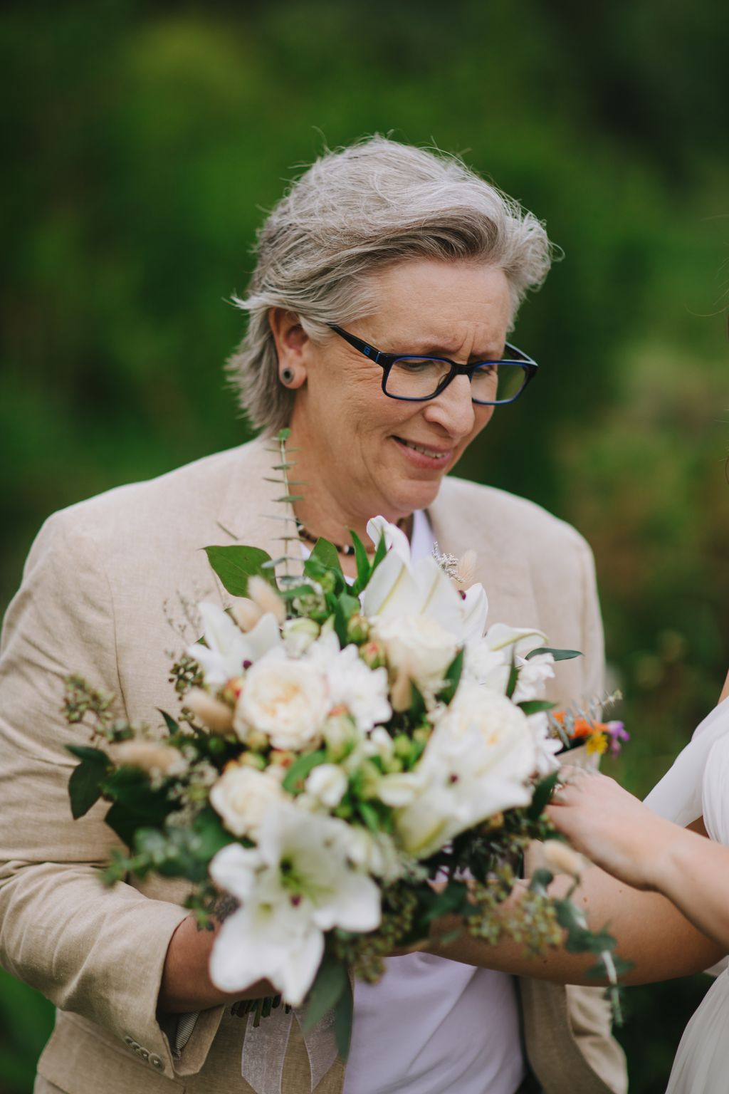 Woman with gray hair and glasses holding white flower bouquet, smiling outdoors with soft green background