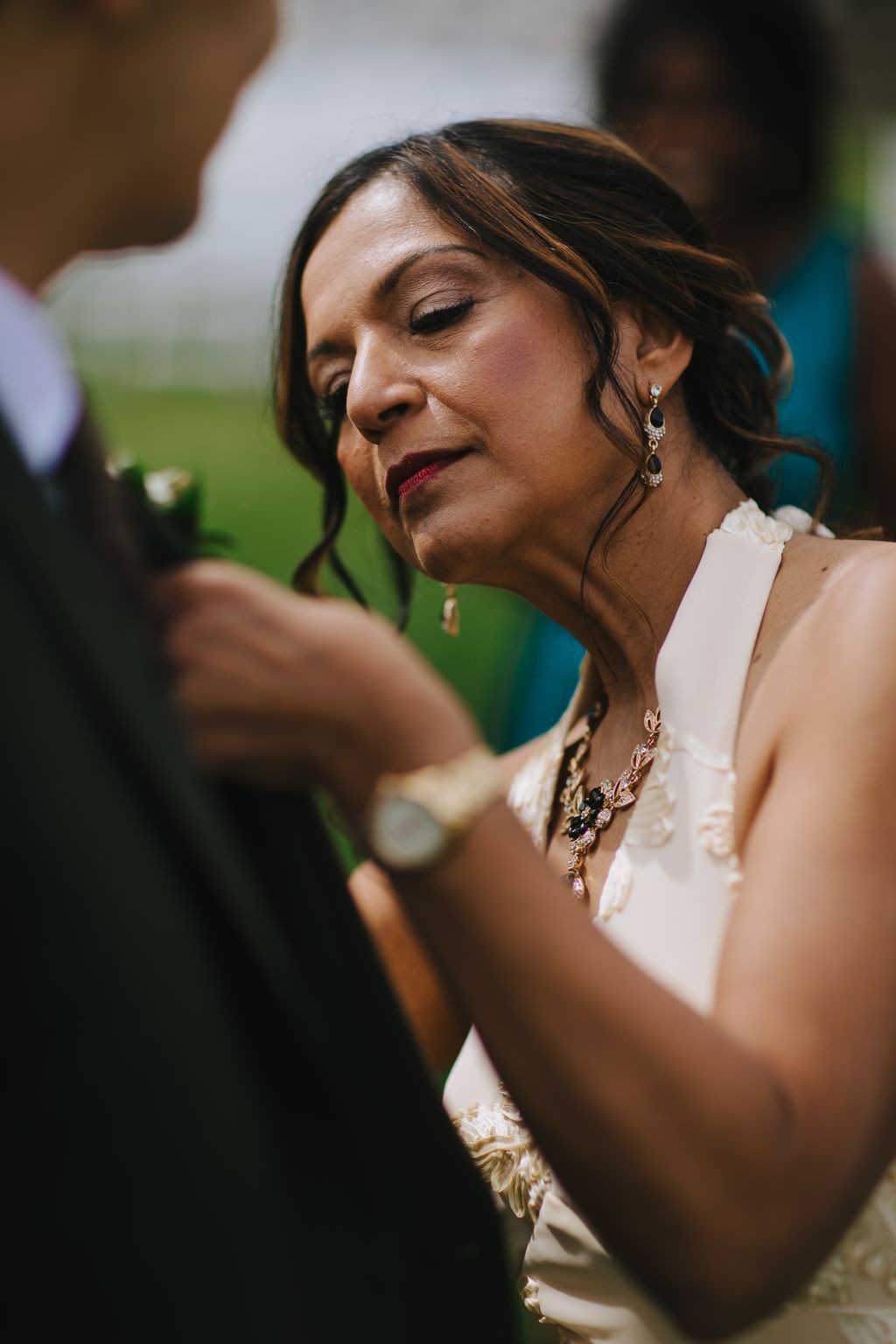 Bride with dark hair and red lips wearing ornate jewelry gazes downward in soft natural light against a blurred green background