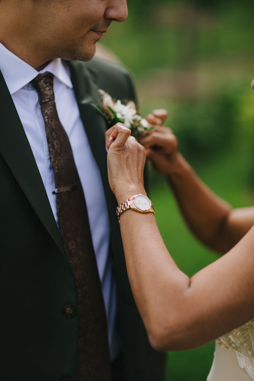Groom in dark suit adjusts boutonniere while wearing rose gold watch, soft green background blur