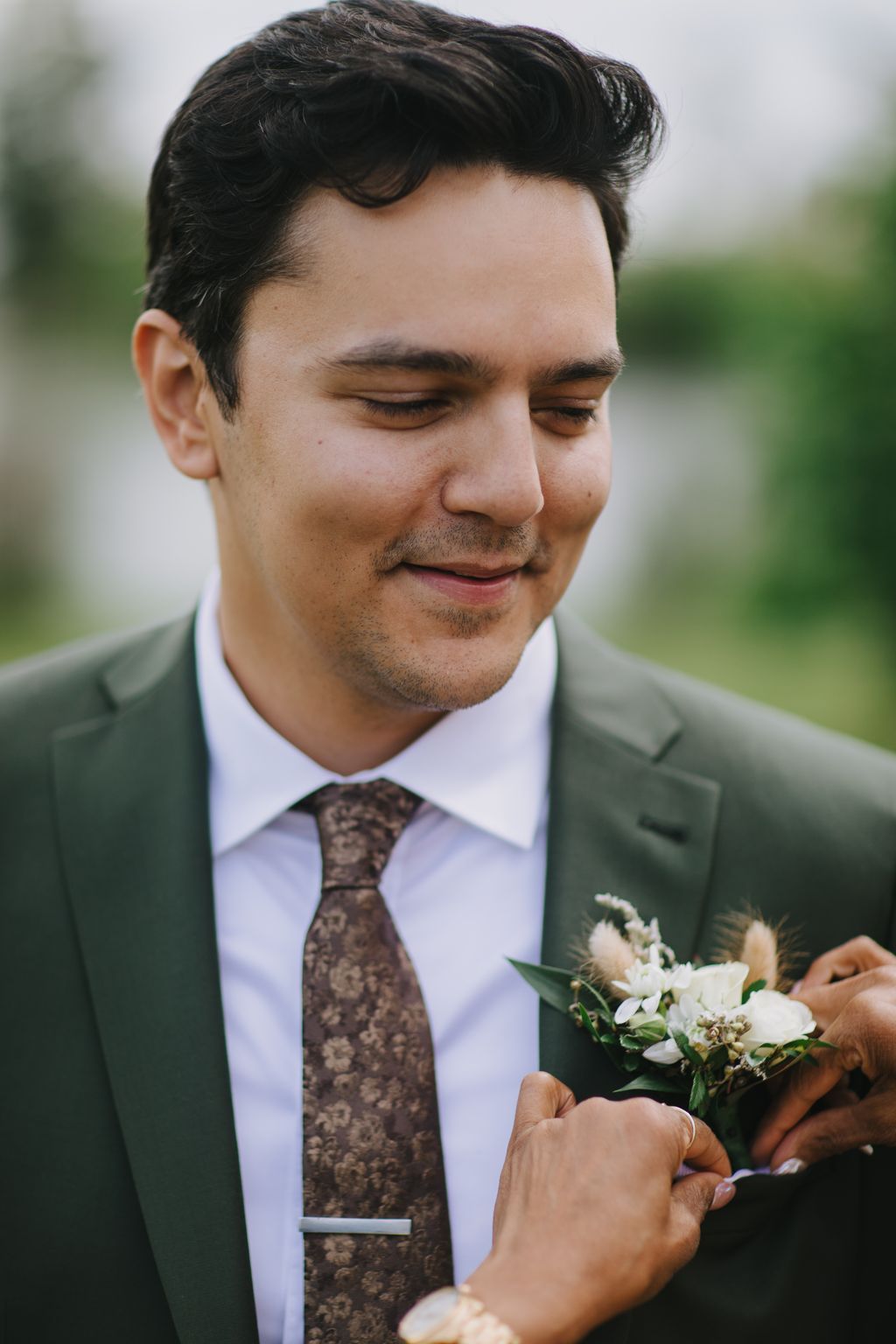Groom in light blue shirt and patterned brown tie gazes downward at boutonniere with gentle smile outdoors