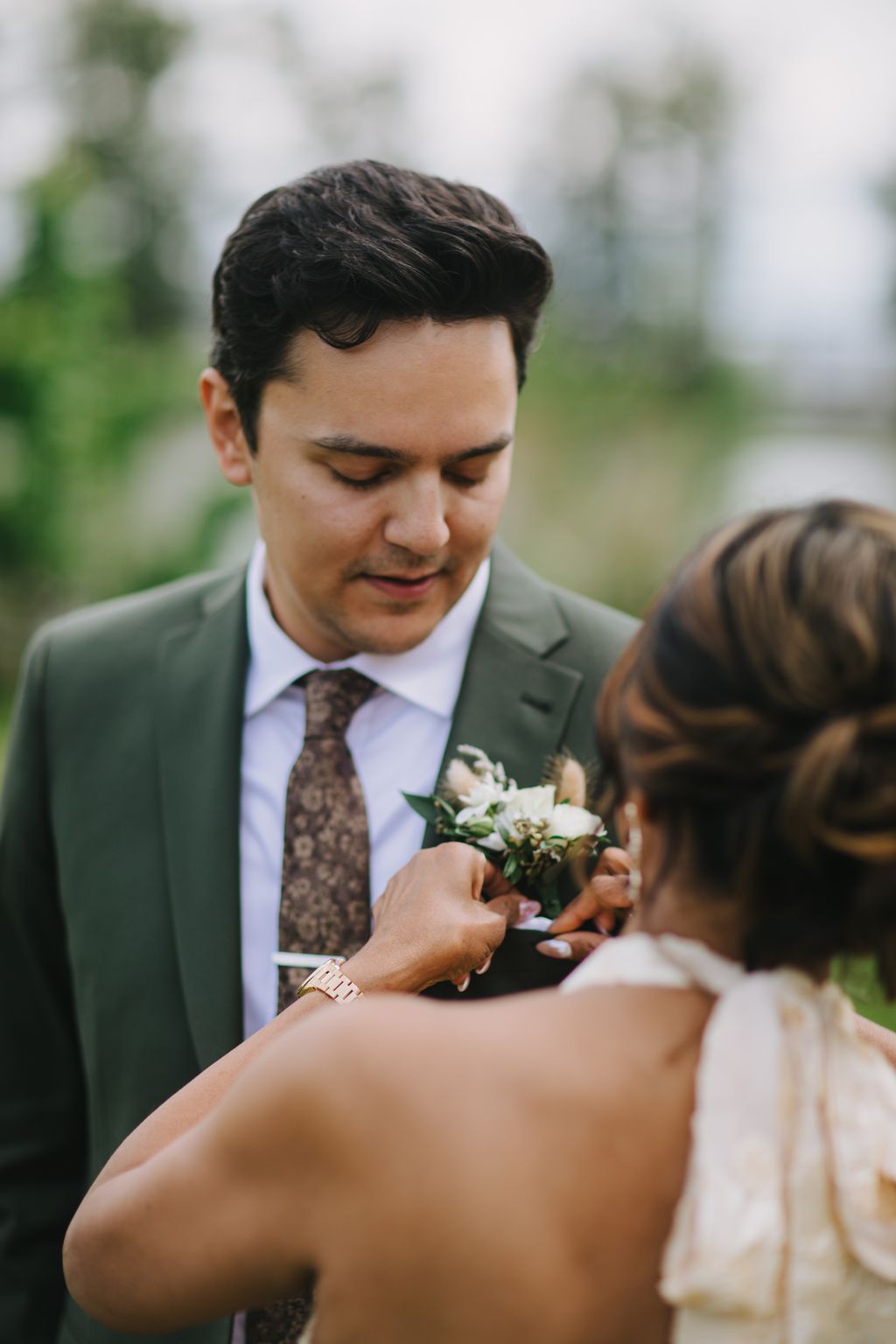 Groom in gray suit and patterned tie gazes down at small white boutonniere held between couple''s hands outdoors