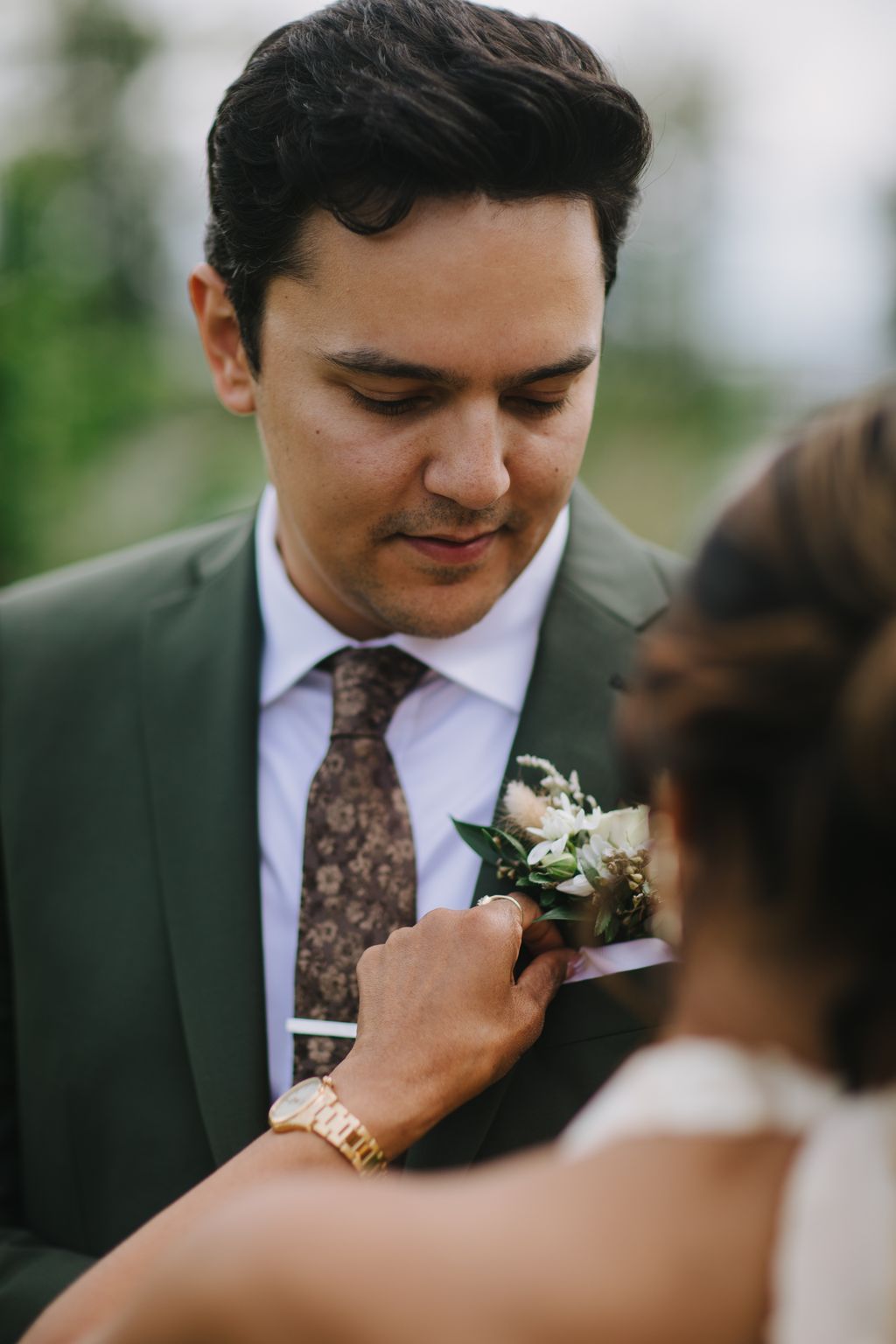 Groom in sage green suit and patterned tie adjusts white floral boutonniere, eyes downcast in tender moment