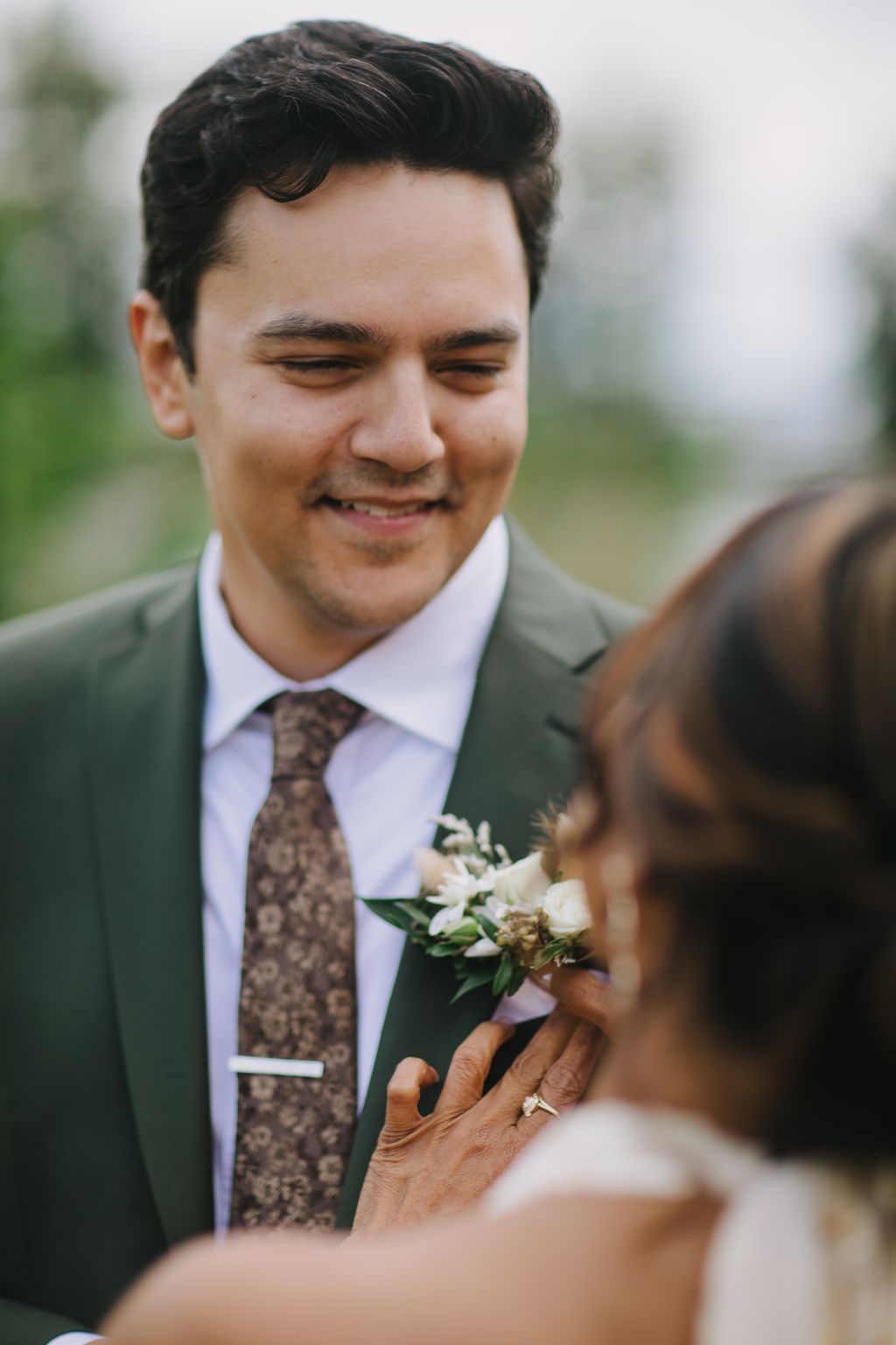 Groom in gray suit and patterned tie smiles warmly while bride adjusts his boutonniere outdoors