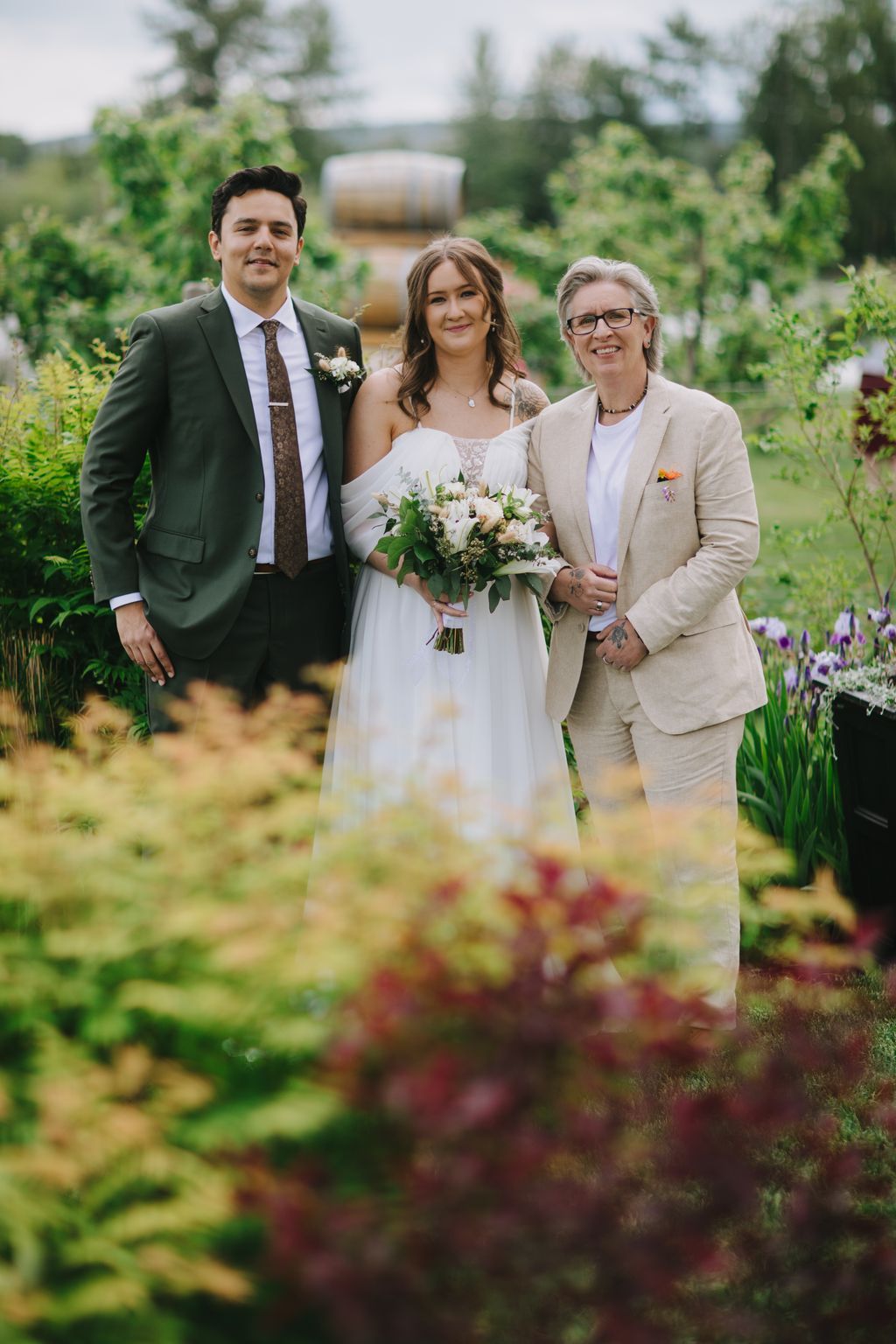 Bride in white dress holding white bouquet stands between groom in green suit and woman in beige blazer in lush garden setting
