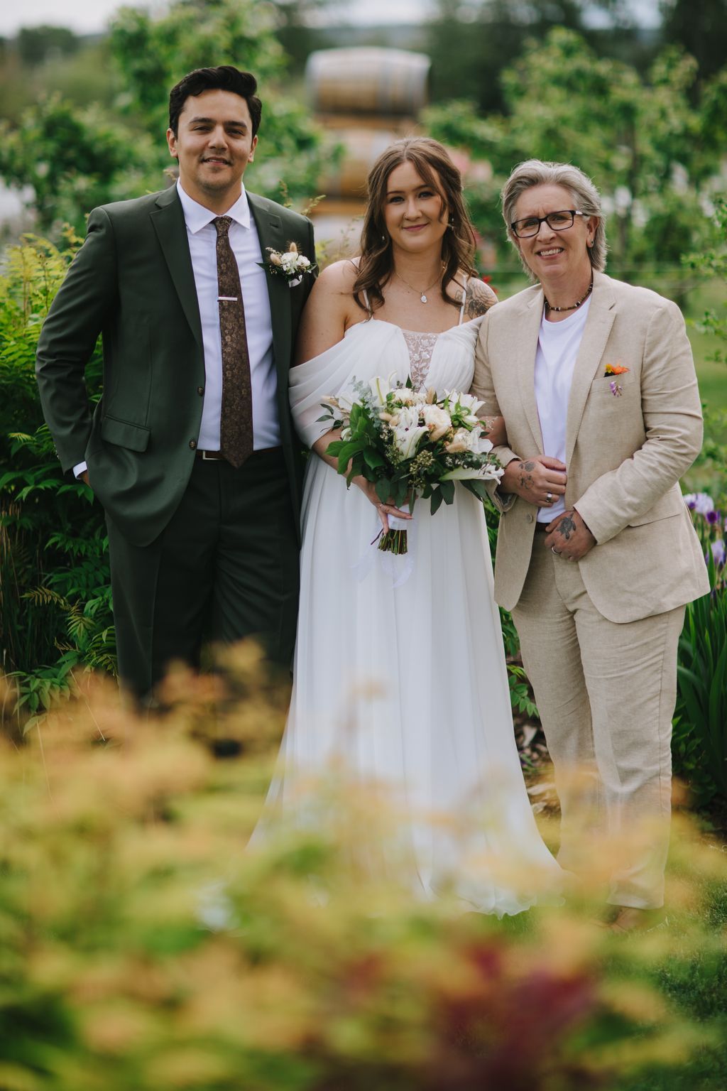 Bride in white dress holding bouquet stands between groom in dark suit and woman in beige jacket in lush garden setting