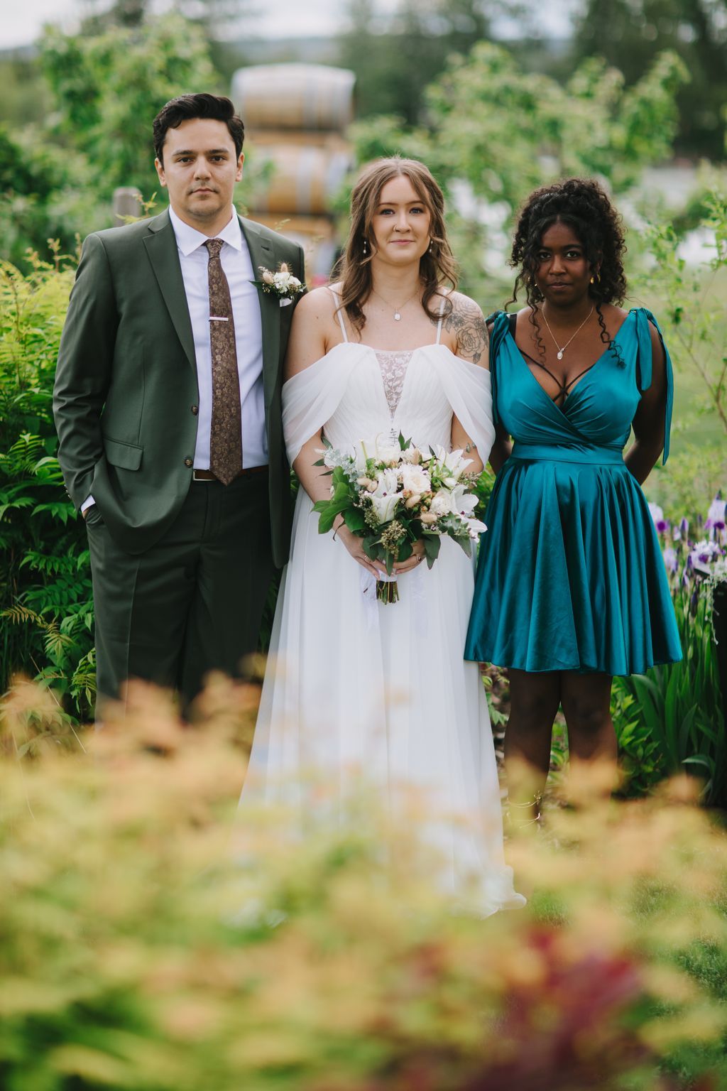 Bride in flowing white off-shoulder gown holds white bouquet beside groom in green suit at outdoor garden ceremony