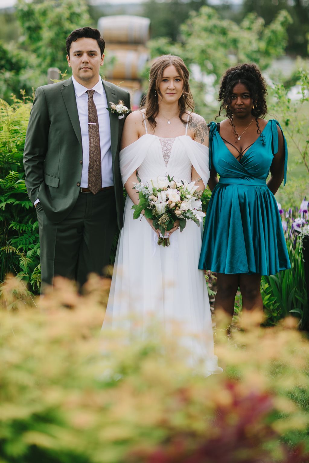 Bride in white off-shoulder gown holding white bouquet stands between groomsman and bridesmaid in garden ceremony setting