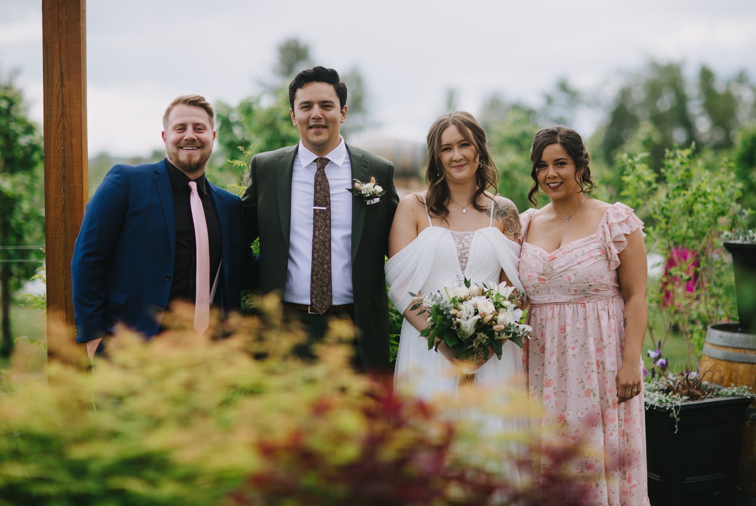 Bride in white dress holding bouquet and groom in green suit with brown tie stand together in sunlit garden setting