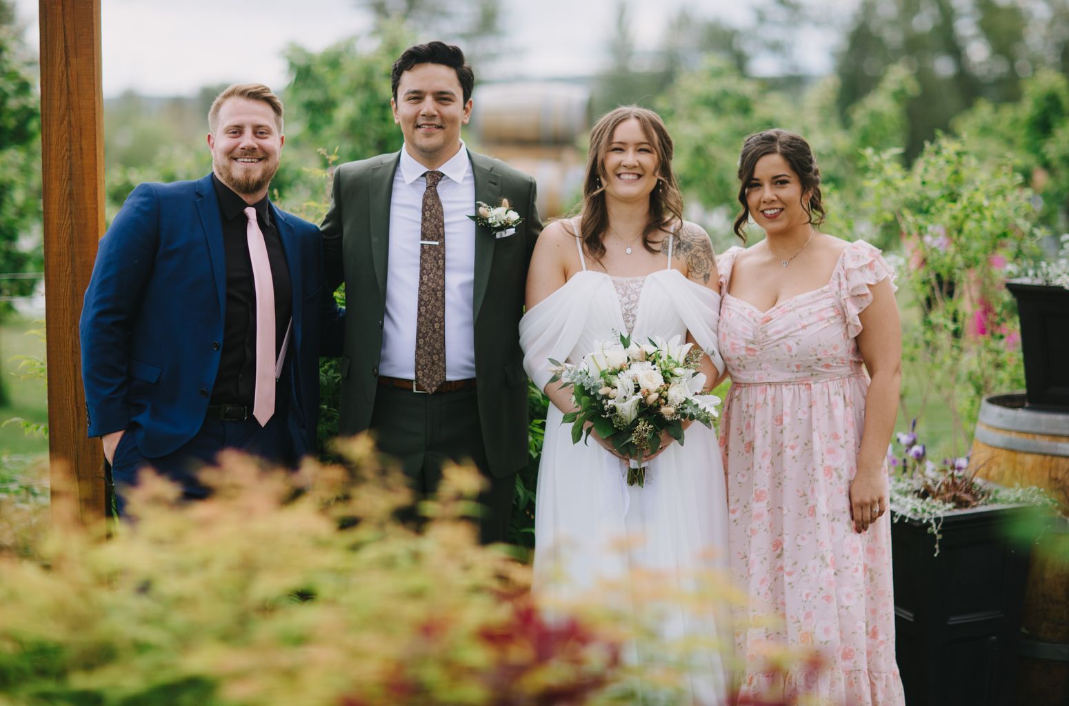 Bride in flowing white dress holding white bouquet stands smiling with groom in olive suit and patterned tie outdoors