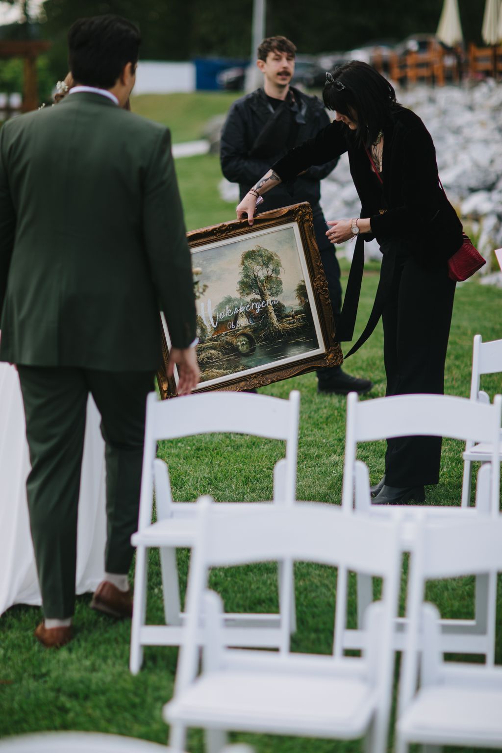 Couple holding ornate framed landscape painting during outdoor ceremony with white chairs and green lawn