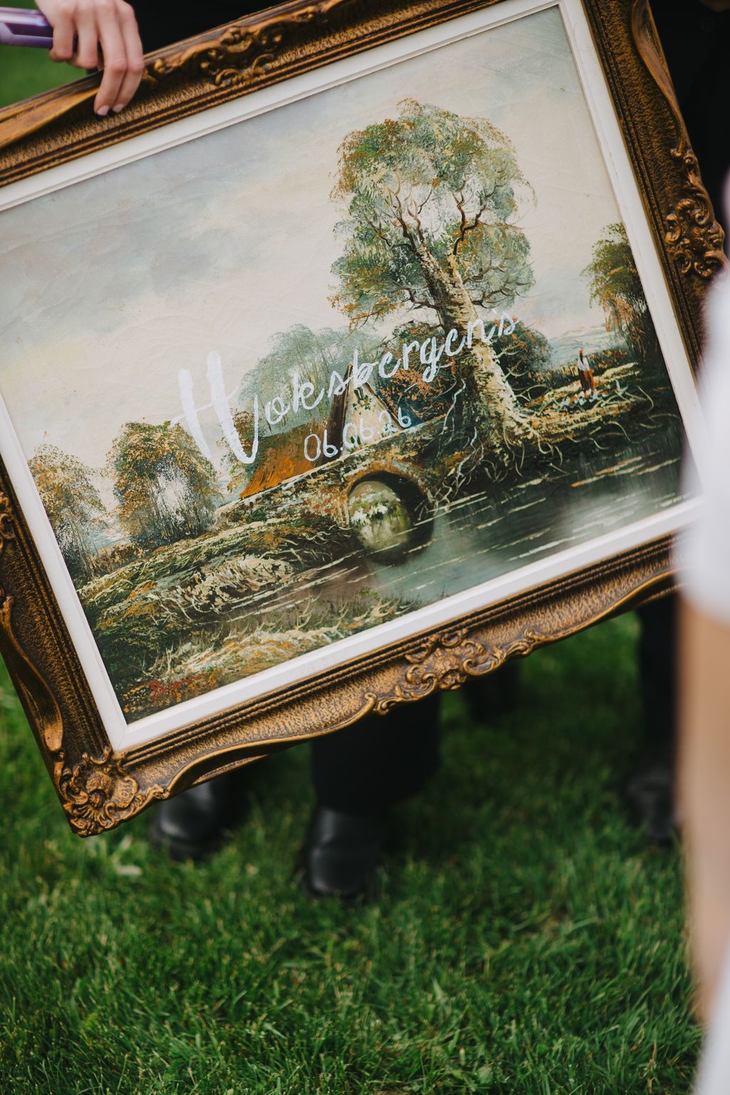 Ornate gold frame containing vintage landscape painting of tree and stone bridge, displayed outdoors on grass