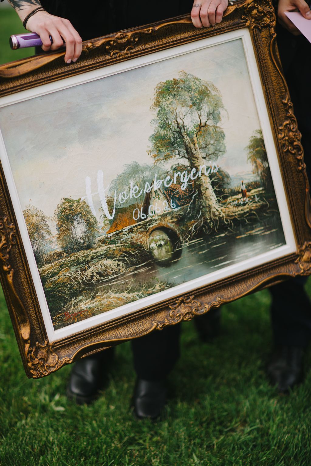 Ornate gold frame holding botanical illustration with visible watermark, held by hands outdoors on grass